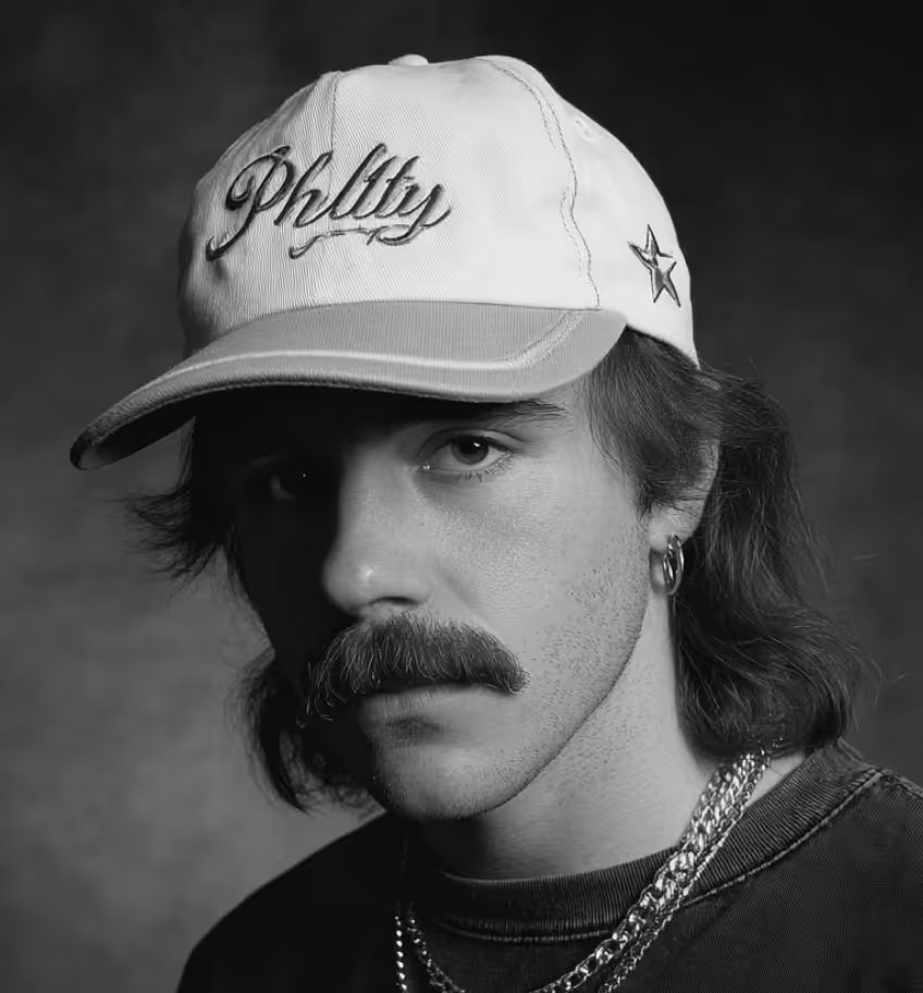 Black and white portrait of a man with a mustache wearing a cap with the word 'Philly', a star on the side, hoop earring, and layered chains.